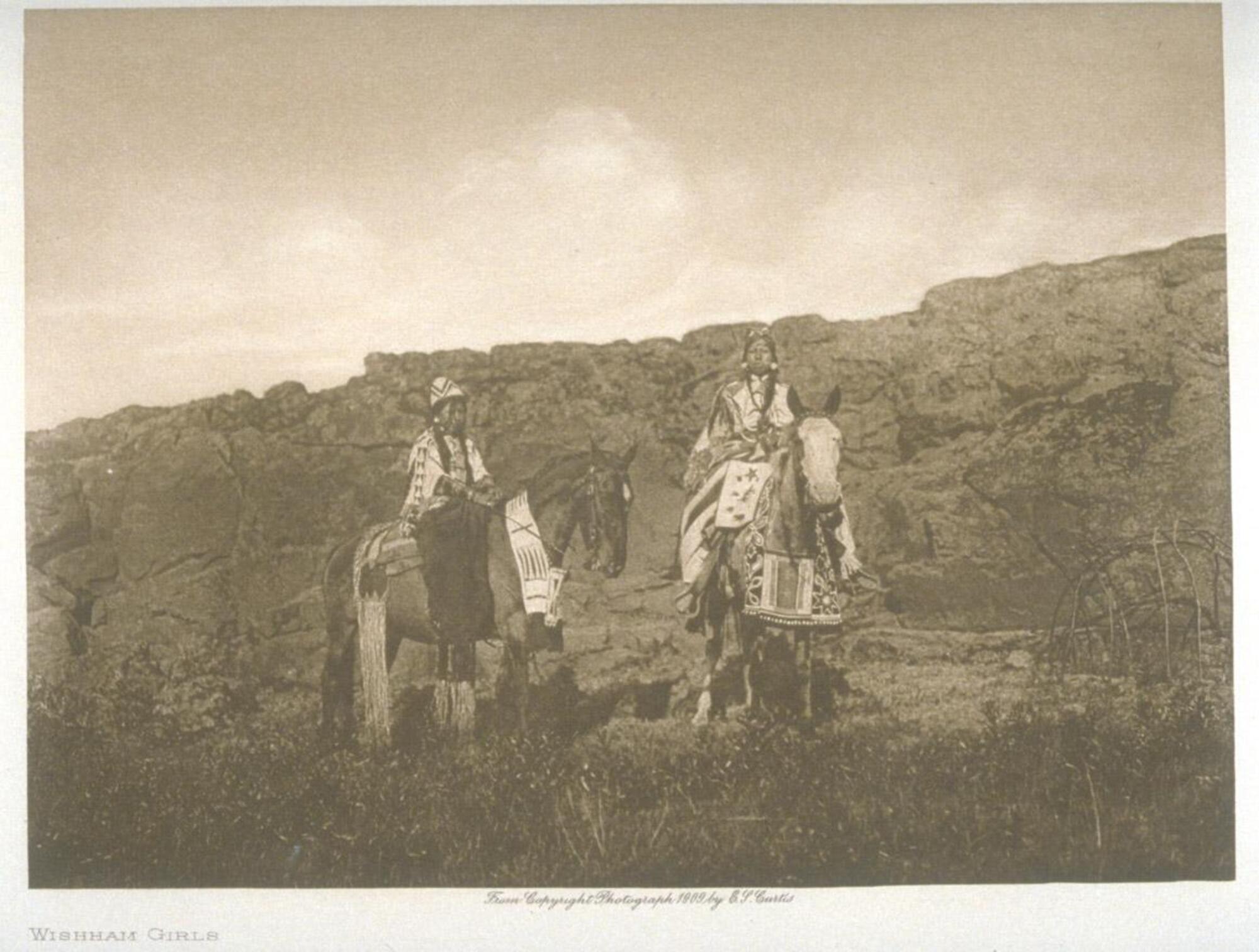 A photograph of two young girls riding horses through a rocky landscape. The girls and horses wear clothing made of fabric, adorned with beadwork and embroidery. 