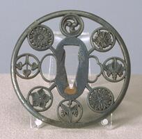 Circular tsuba, made of iron. Inside an exterior circle, eight smaller circles are placed with the same spacing. The eight circles are connected to the exterior circle as well as to the three center holes where kôgai, blade, and kozuka are placed. Each of the eight circles have a different family crests. The openwork technique seen here is called "marubori" (round carving). The surface is slightly textured by minute stippling.
