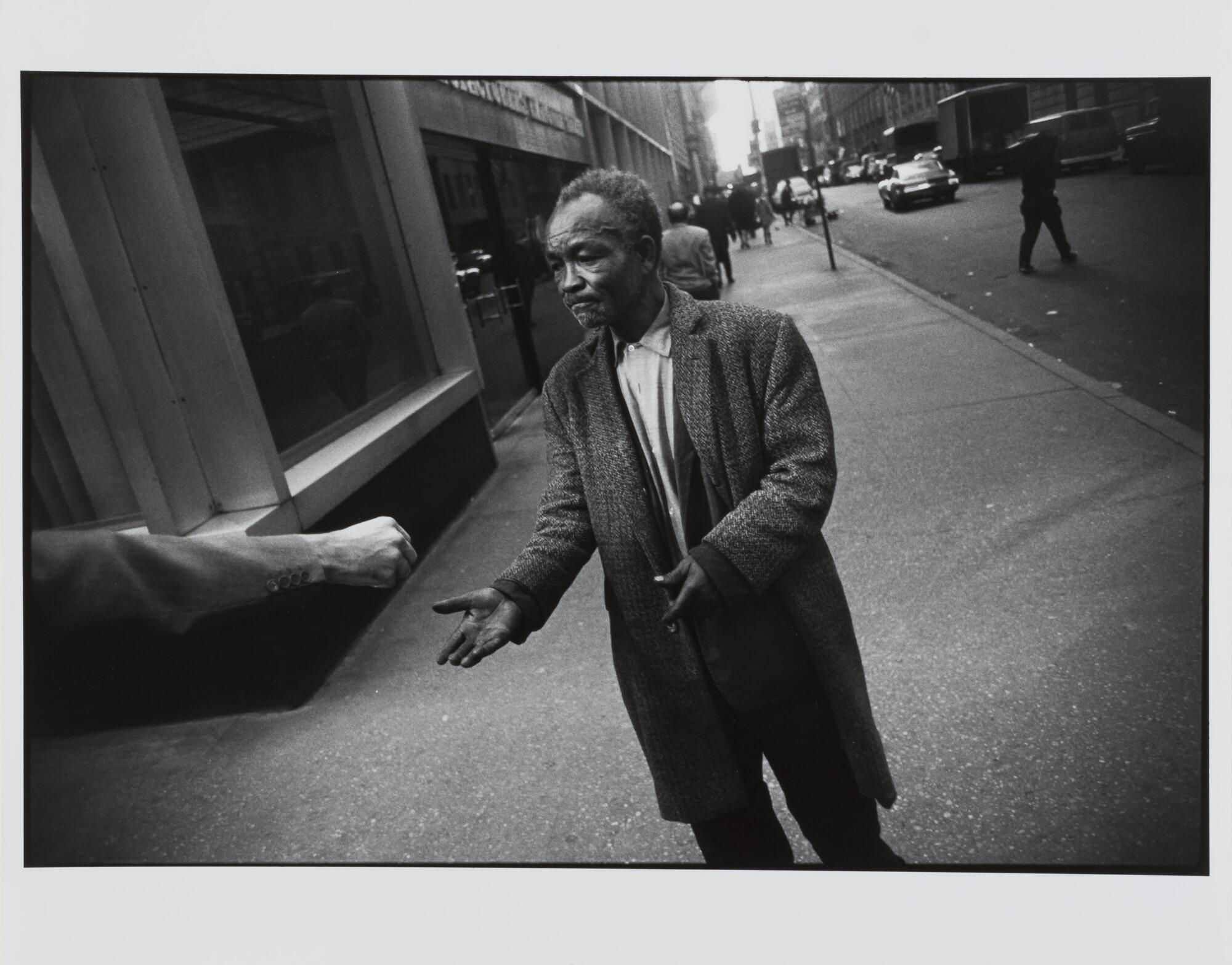 This photograph depicts a view of a man accepting a coin from a passerby on a city sidewalk. 