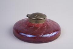 Inkwell made with glass of cranberry red color has a flat around body and a matel lid cover.