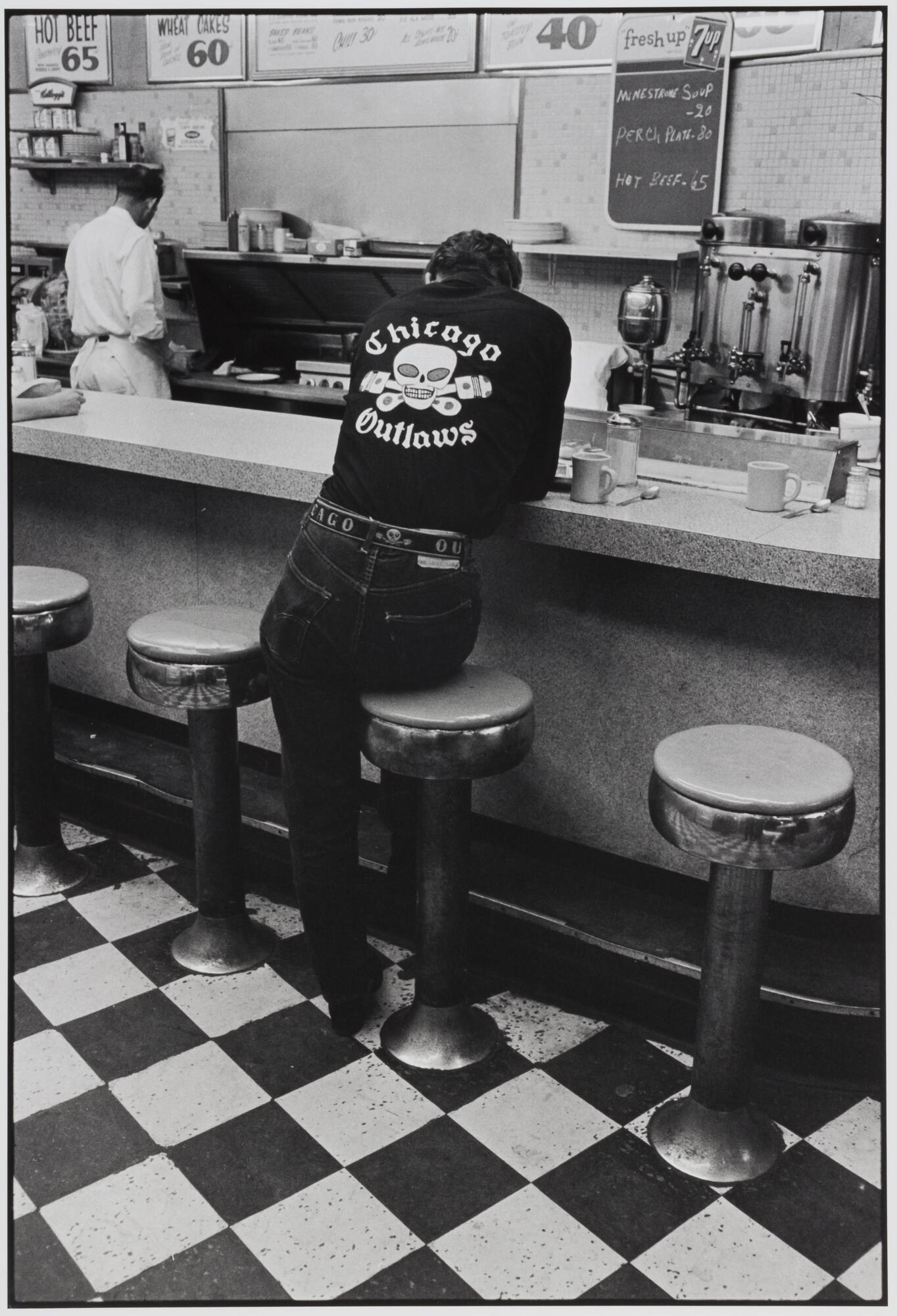 A photograph of a man sitting on a circular stool in a diner, his back turned to the camera, revealing a shirt, which reads "Chicago Outlaws." Behind the counter, a man prepares orders. 