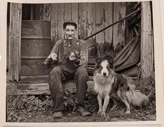 A man holding a violin sits on the porch of a wooden structure. A dog stands to the right of the man.&nbsp;