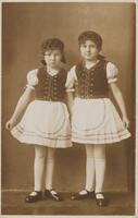 Two children standing next to each other and holding their skirts.