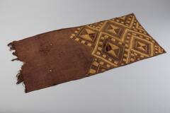 Rectangled panel with hemmed edges. One side is frayed and not hemmed. There are four large diamond designs partially covering the panel consisting of brown diamond shapes, tan chevrons and a linear checkered tan and brown pattern. There are four small holes on the undesigned brown surface. Additonally, there is a modern repair panel attached to back of the fabric.