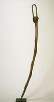 This curved wooden Pende staff features a finial depicting a human head bearing simple facial features and a cap-like coiffure composed of vertical lines. The staff’s handle is in the shape of a narrow loop that connects the front of the figure’s head to the back of its head. A slender serpent carved with a snake-skin pattern slithers upwards from the lower end of the staff.