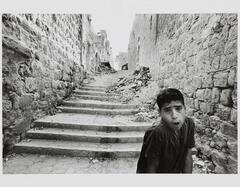 Image of a dark-haired boy with open mouth in a rubble-strewn stairway between buildings. Far in the background, a woman watches.