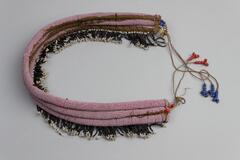 Beaded necklace made up of three rounded attached pieces of material with pink beaded overlay. White and black beaded fringe on the inside. Tie closure with strings coming off either end with red and blue beaded ends.