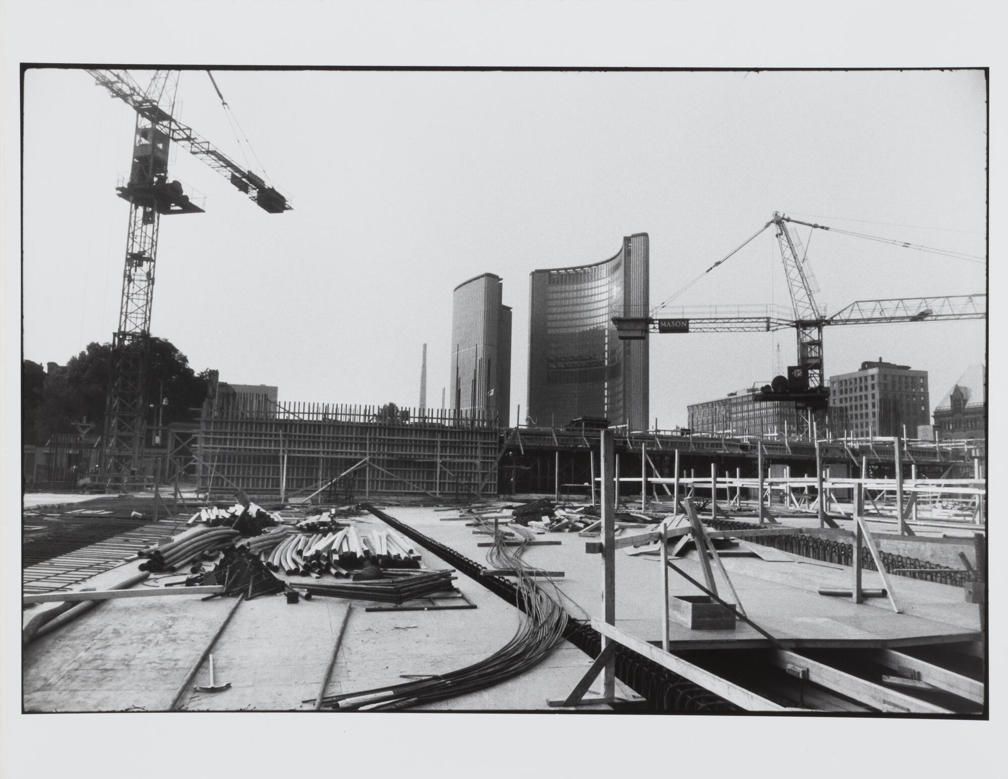 This photograph depicts a view of the construction site of a large building. There are piles of materials, two towering cranes and a view of other buildings in the background.