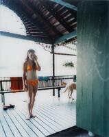 This photograph depicts a young girl in a two-piece bathing suit standing on a porch and looking into an open doorway. Behind her is a dog and a body of water. This image is the far left panel of a triptych.