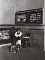A photograph of a shoeshiner sitting outside of a bank decorated with large signs. He sits in a corner between the building and a subway entrance with his equipment in front of him.