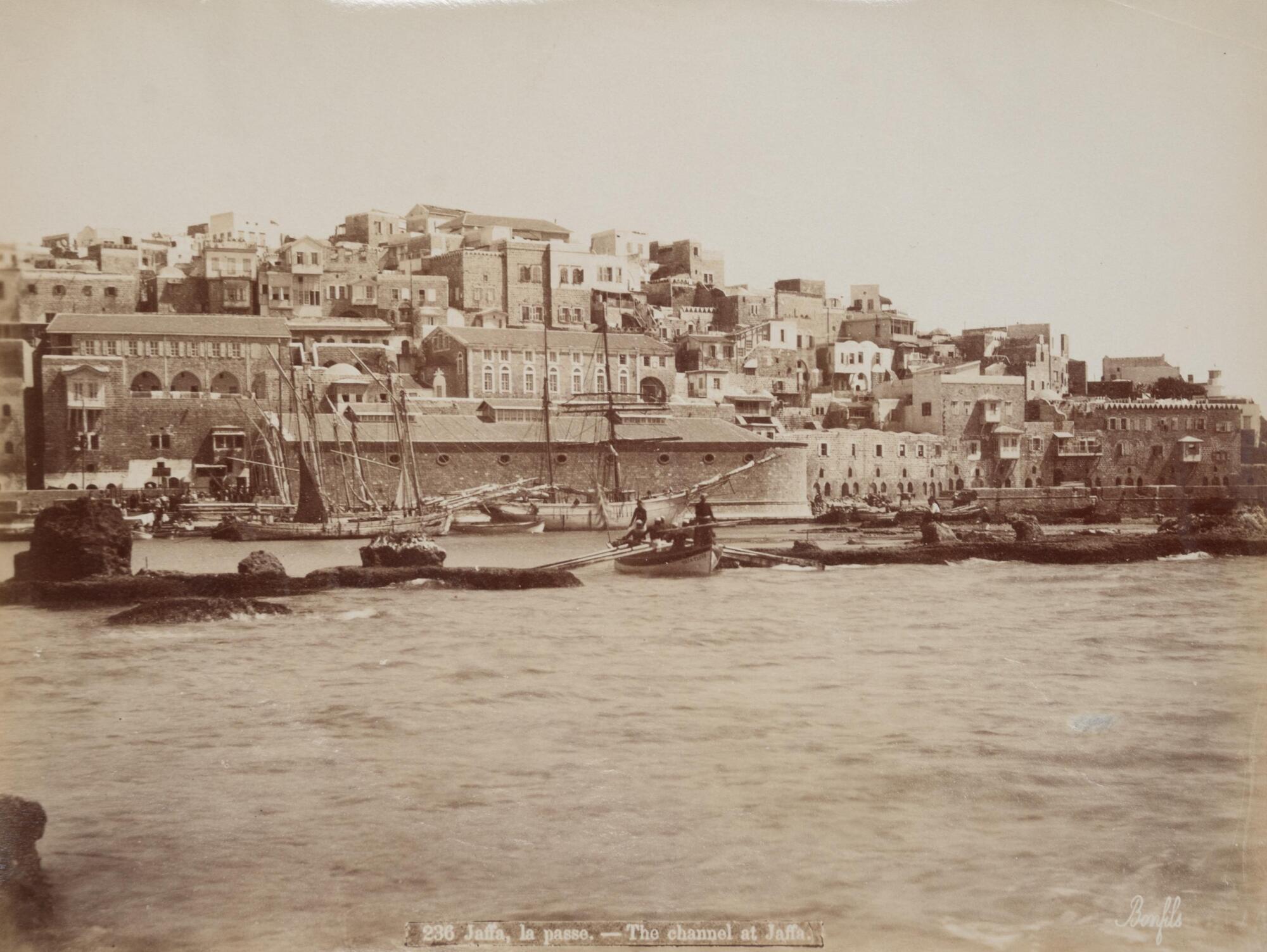 View of a wharf from across a channel. Ships and small boats populate the middle ground, while a city rises behind. The foreground is mainly seawater.