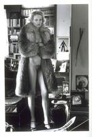 A photograph of a glamorous woman standing in her home, wearing only a fur coat, necklaces and heels. 