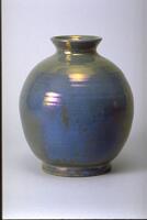 Footed vessel with round body, short neck and flared mouth covered with an iridescent glaze over a semi-matte glaze that creates an appearance of irregular patches of color. It has an overall blue-green appearance. The rings of the thrown clay can be seen beneath the glaze.