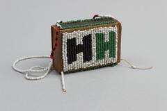 Beaded wooden box with the sides open. Two longer edges with beaded trim in green. Two larger facew with beaded detail. One face in white beads with beaded rectangular detail in blue and red. Other face in white beads with two "H" shapes in black and green. Long beaded handles, white tag attached.