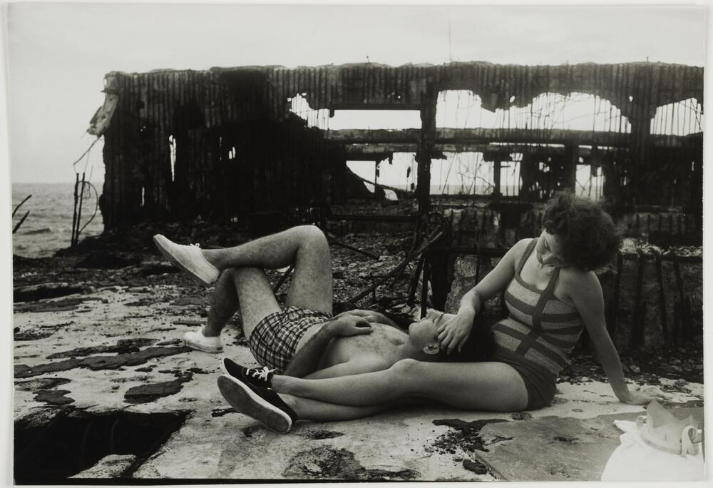 A man and woman in swimwear lounge on a surface of decaying manmade materials. Behind them stands the exposed skeleton of an abandoned structure. Beyond that, a body of water stretches to the horizon.