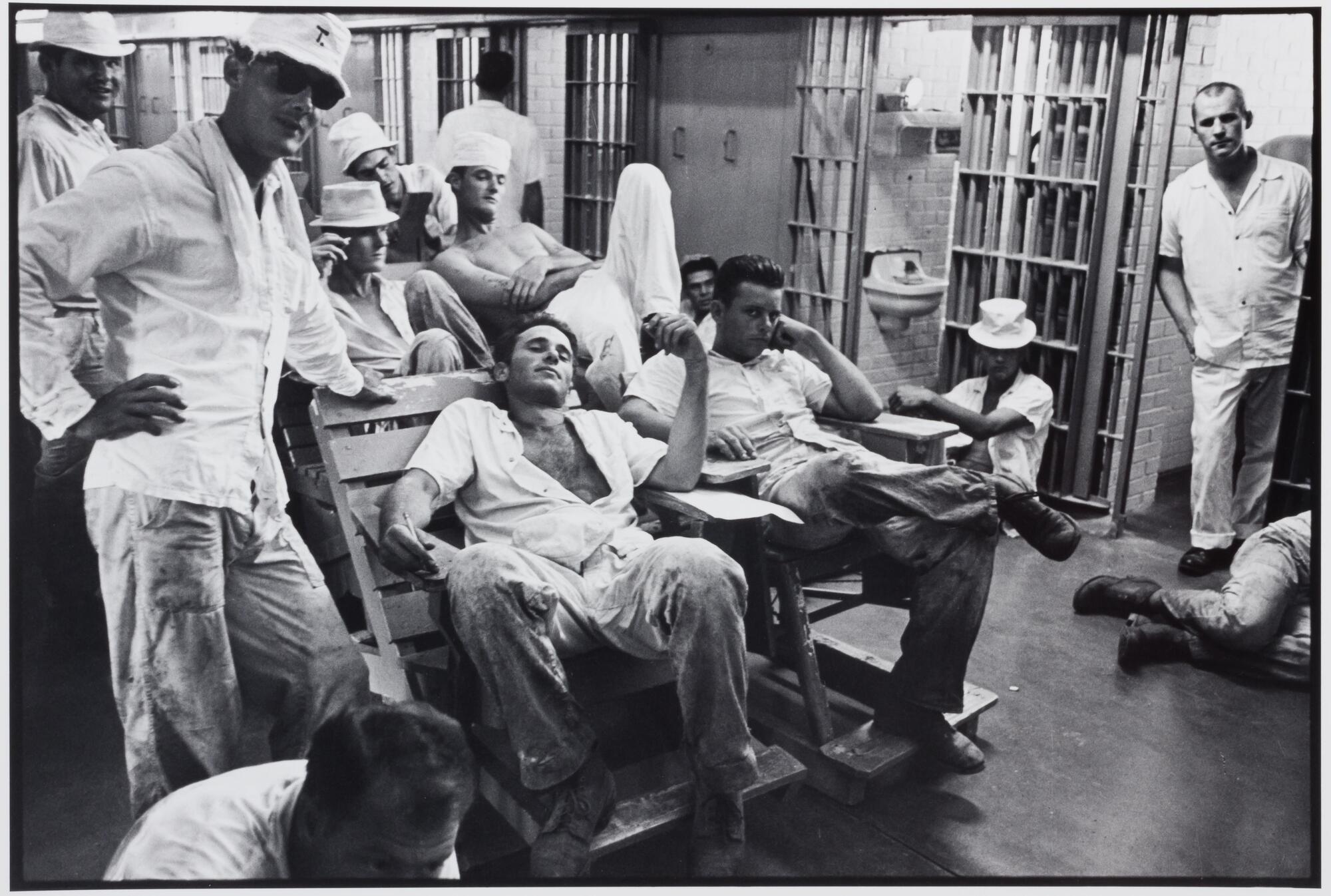 A group of prisoners in a relaxed area of a cell block. Most are seated and some are standing.