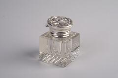 A square, crystal inkwell with a silver lid. The lid has a carving of a woman's face and hair on the top.
