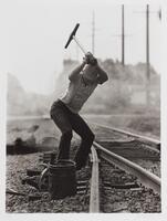 Railroad with a man swinging a tool over his head. There is a bucket to the side of him, telephone poles behind.