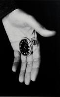 A black and white photo of a hand holding a small pocketwatch. The hand appears to be letting go of the watch.