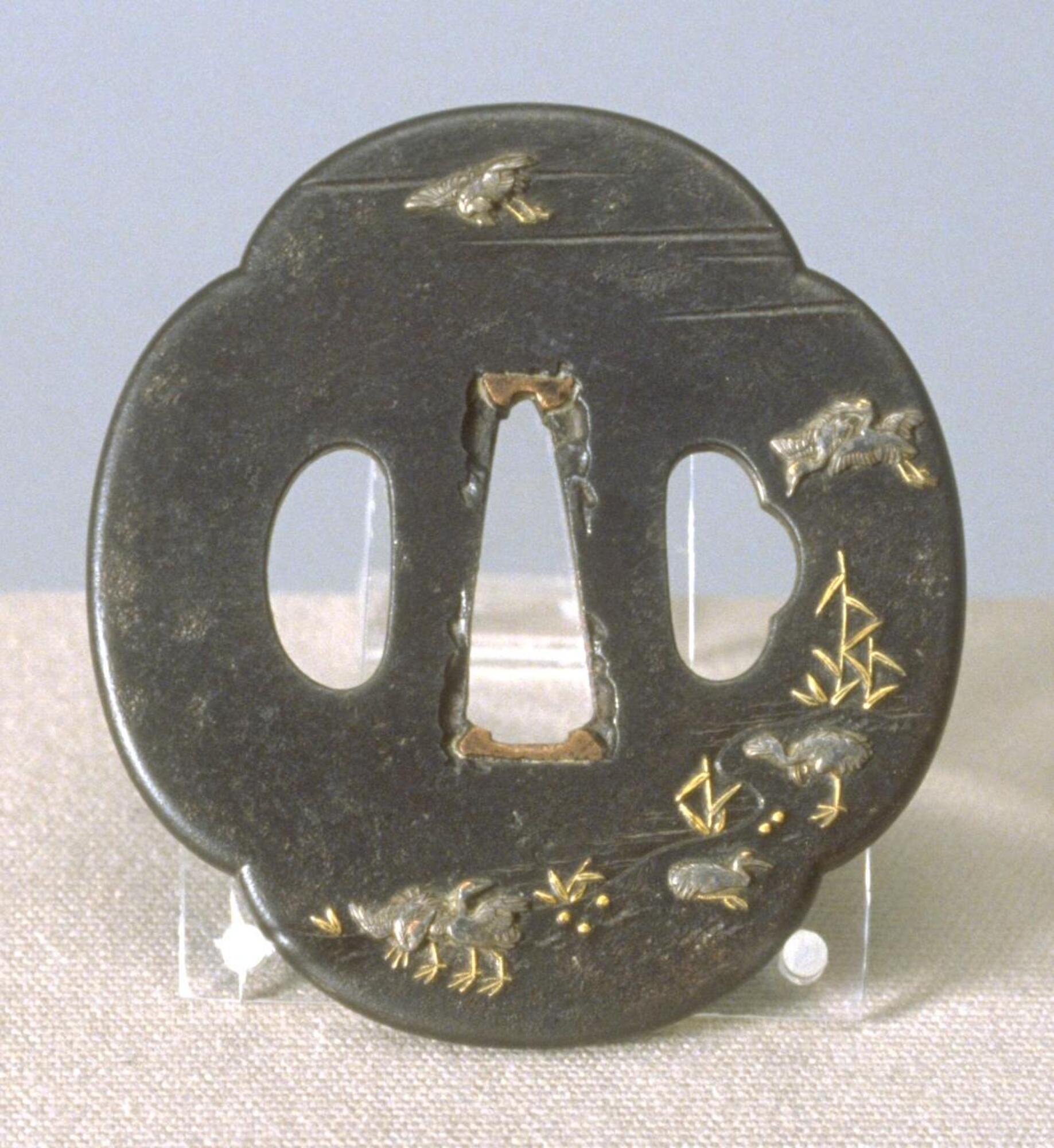 This tsuba is a flat iron plate with quatrefoil design. It has three holes: one for blade (middle) flanked by oval-shape hole (for kougai) and oval with bump shape (for kozuka).  Egrets and reeds decorate the surface, distributed in a curve that climbs counter-clockwise from the bottom left register, culmiating in the top left with a lone egret in flight.  Egrets on the bottom of the piece perch on the ground or nest in the golden reeds.<br />