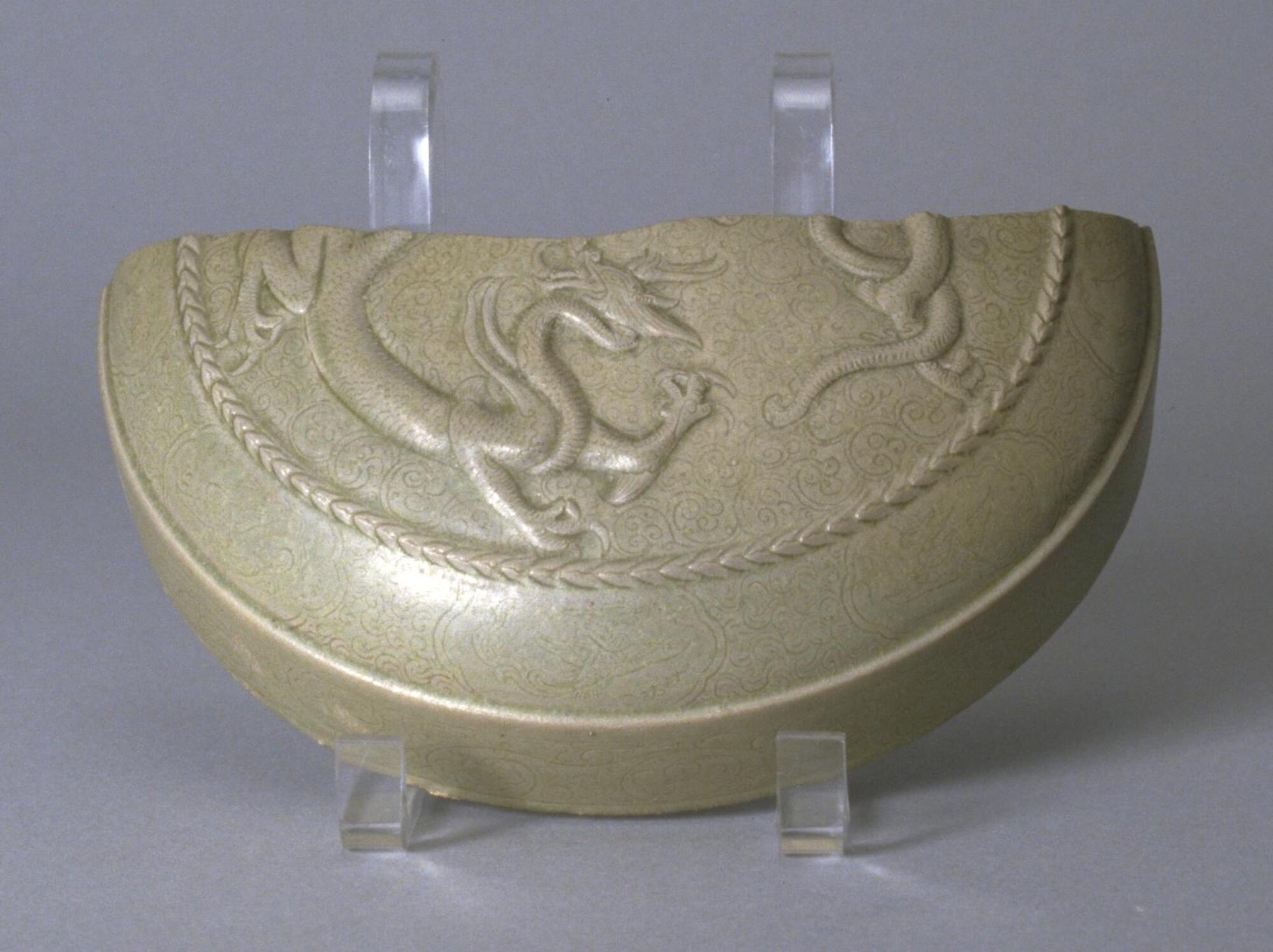 This fragment of a stoneware box lid has straight sides with a shoulder curving to a flat top. The lid has a finely detailed dragon in relief inside a twisted rope roundel border against a ground of finely incised floral meander, covered in a light gray celadon glaze. 