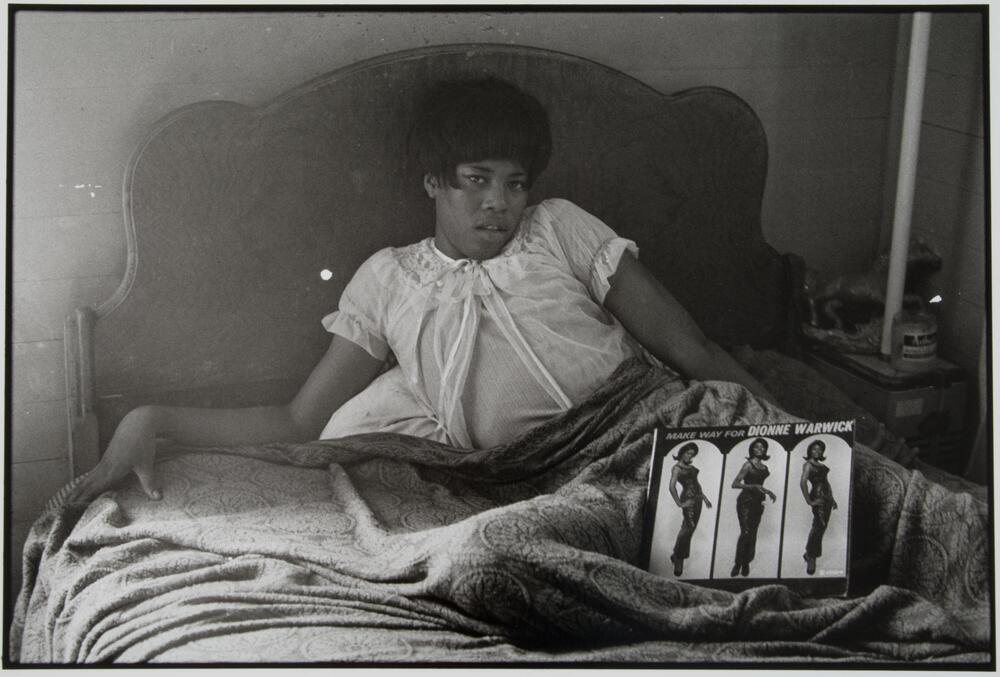 A photograph of a woman reclining in her bed. A vinyl record titled "Make Way for Dionne Warwick" is propped up on her legs.