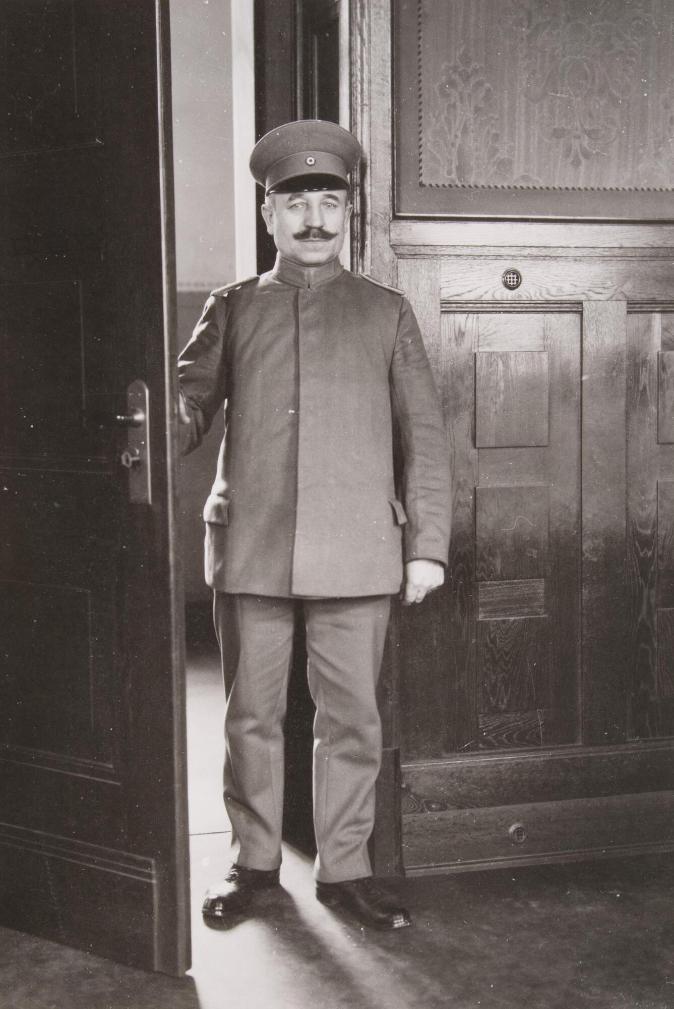 This photograph show a man standing inside of a court house. He is fully dressed in a uniform, standing in a doorway.