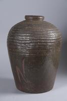 A vase made of slab on wheel with long bottom and a circular opening at the top. Dark brown with ribbing/striped pattern on the upper half, chatter marks on upper 3"; rolled lip and a greenish brown semi-gloss glaze.