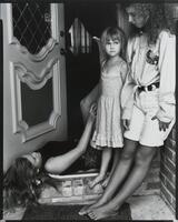 Three girls in a doorway. A little girl in a floral dress is standing, an older girl is lying down and the third, is standing against the frame.