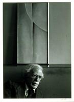 This photograph shows the photographer Alfred Stieglitz seated in an interior, a tall, vertical painting by Georgia O’Keeffe positioned on a picture railing above his head.