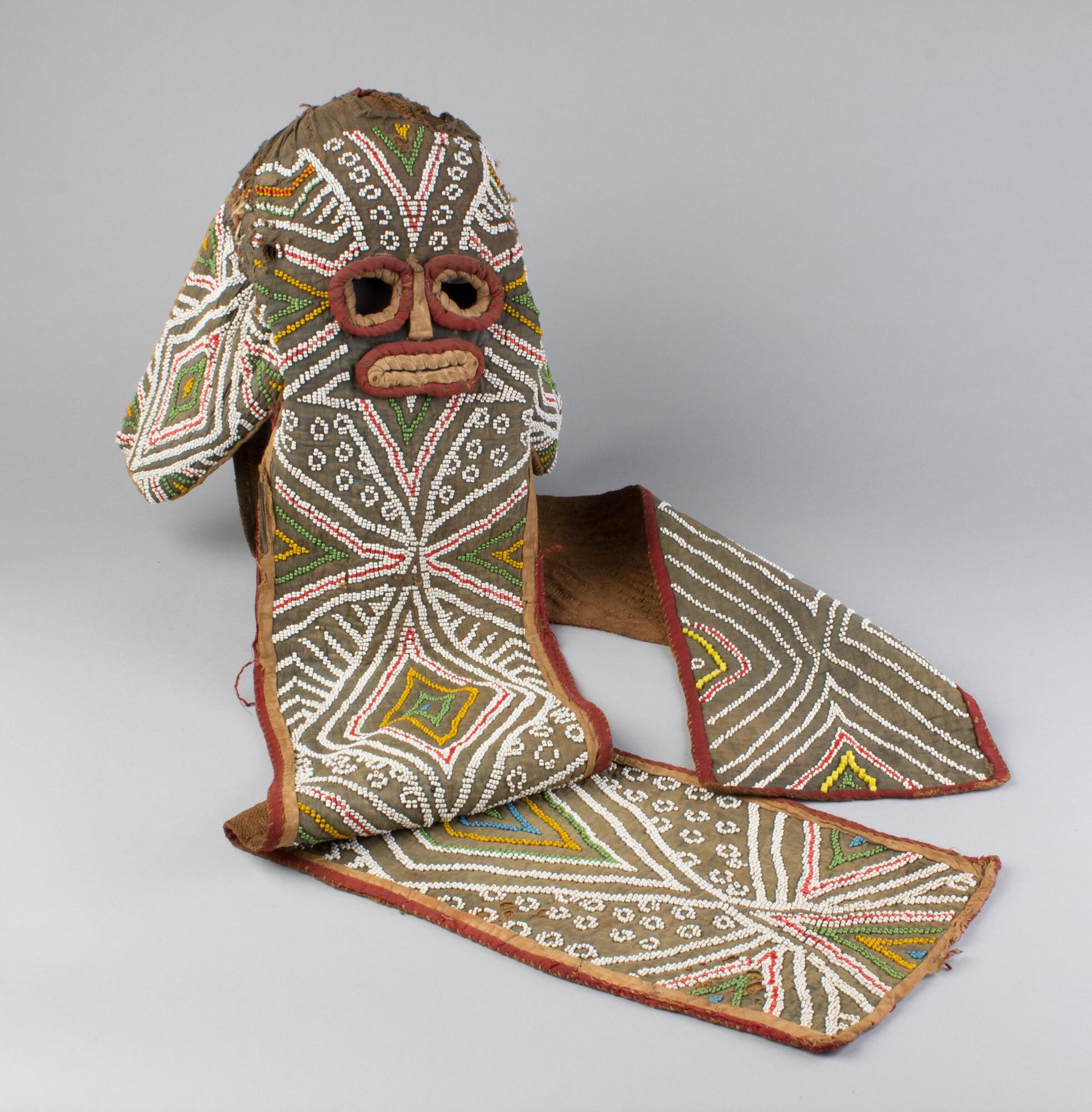 A fabric mask with round eyes and an oblong mouth and nose made from red and yellow bound cloth. The brown base fabric is covered with small beads in a geometric pattern. The majority of the beads are white, with red, yellow, green, and blue beads also used. The edges of the mask are bound in red and tan cloth. 