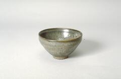 A conical bowl on a tall straight foot ring, covered in a thick dark brown-black glaze with gray mottling. The too-thick glaze is crawling away from the underlying clay body, with a fine crackle covering the glazed surfaces. <br /><br />
9/8/16, RCE: Minor changes to syntax