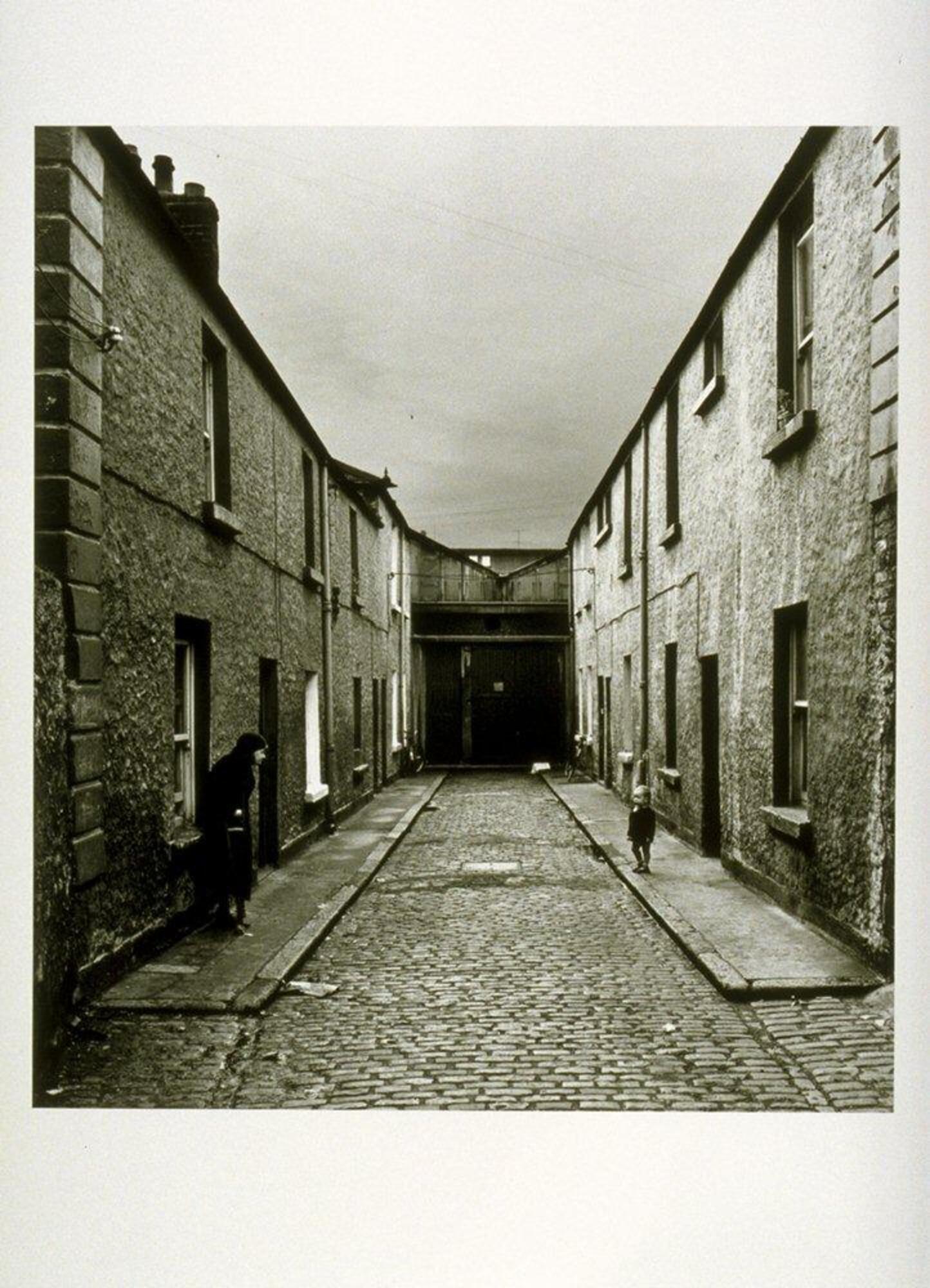 This photograph depicts a view of a cobblestone alleyway. On opposite sides of the street stand an old woman and a young boy.