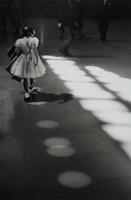 A girl in a short dress walking next to the light coming in from the windows. Man walking towards her, she has a very dark shadow.