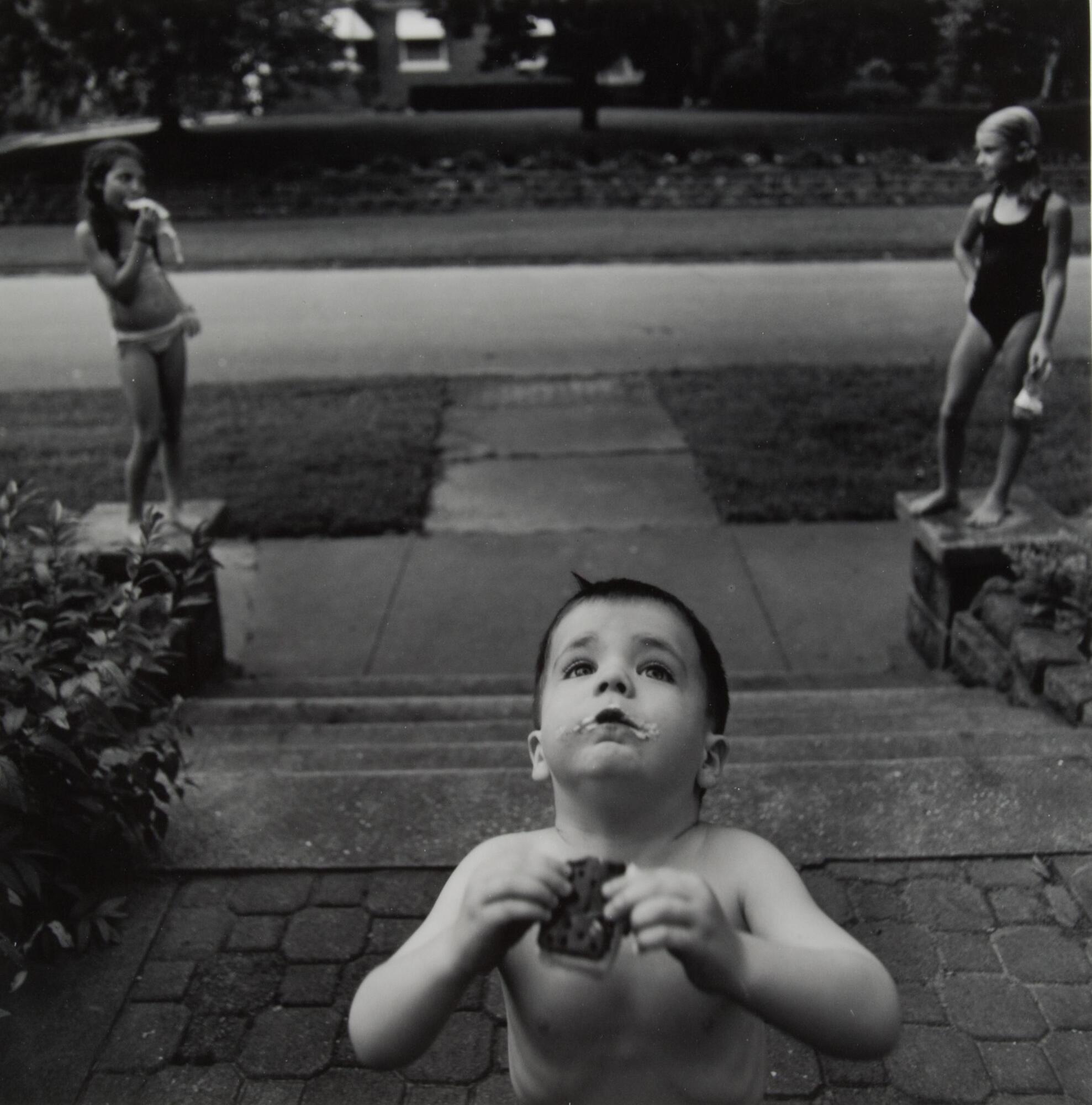A photograph of three children in front of a house. Taken from inside, looking out; a young boy eats an ice cream sandwich in the foreground. Two young girls stand on square columns at the end of the steps leading to the house, framing the boy and mirroring one another's pose.