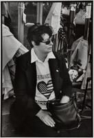 A woman seated wearing a jacket and a memorial tshirt. She has on sunglasses and is looking to the side. There are people on either side of her.