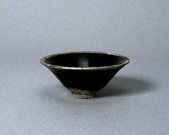 A conical stoneware bowl with an everted rim on a tall, straight foot ring.  It is covered in a dark brown-black glaze, with an unglazed rim and several chips to the rim.