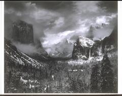 The photograph depicts a wide view of a mountainous valley in winter with a storm sweeping through its interior in the distance. To the right, a waterfall spills over a ridge.