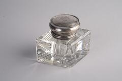 A small. square, crystal inkwell with a sterling silver lid. A star is engraved on the bottom of the base. The lid is monogrammed.