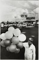 A smiling young man in light coveralls holds a bunch of twelve balloons in the foreground of this image. Far in the background, several cars are parked at a busy filling station.