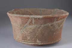 A tall ceramic bowl with a flared mouth. A flat bottom sits on a narrow base and a 90 degree angle creates the bowl itself. Rough red matte ceramic, no glaze. Spattered lines of lighter tan color.