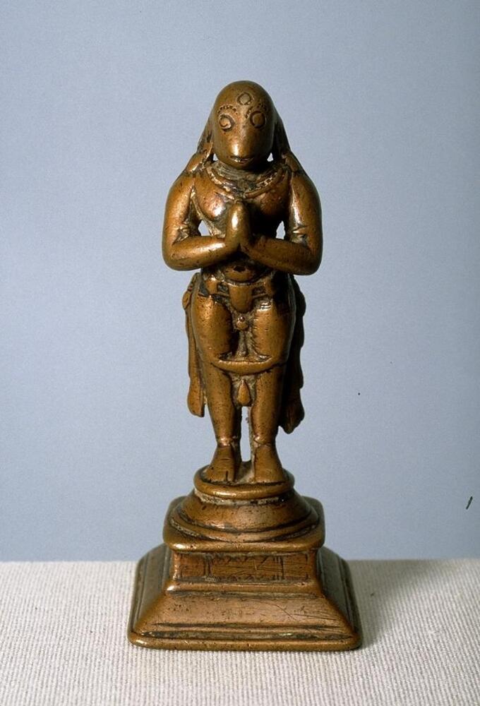 Hanuman stands on a tiered base, which starts square and sloped, then is square and straight up and then round sloped and ringed.  He has a human body and a monkey’s head.  He stands in an unbending stance with his hands in anjali mudra, held up to his chest with palms together in a prayer gesture.  He wears a diaphanous garment from the waist down with only the edged decorated and depicted with a series of belts and sashes hanging in front of it.  He wears a number of necklaces and armlets.  His face is very worn and far less realistic than the body with the large round eyes incised with eyebrows above and a large circle on his forehead. His mouth is a simple slit.  <br />