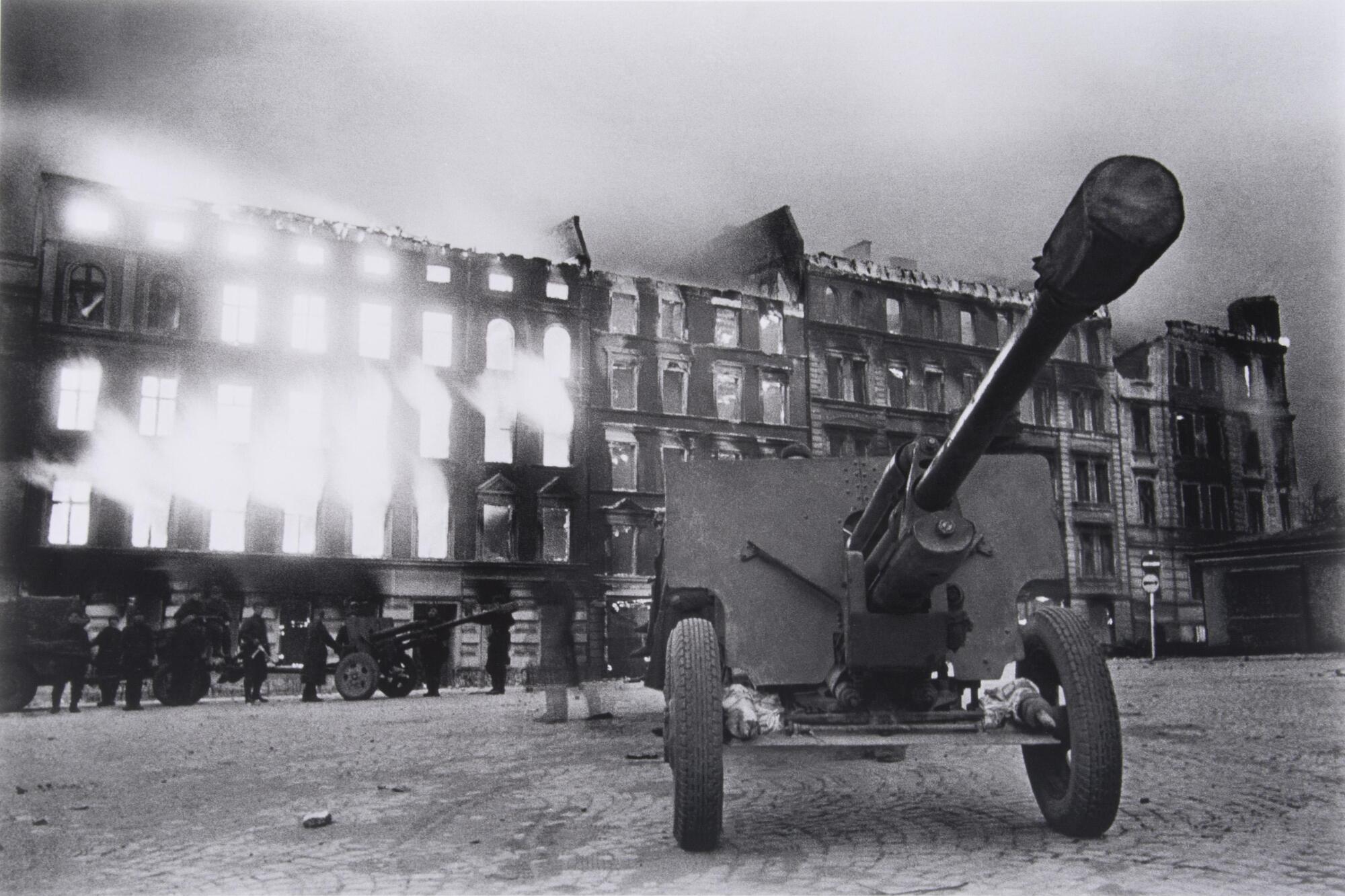 The barrel of an antitank gun points out of the picture plane; a building has caught fire in the distance. 