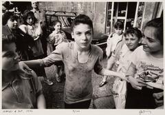 This photograph depicts a group of children in a city street. One young girl stands at center of the group with her arms out. She stares directly into the camera, while the others look at her.