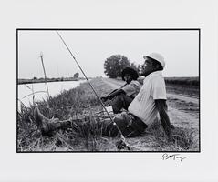 A black-and-white image of a man and woman sitting on a strip of grass which separates the dirt road on the right from the water on the left. A fishing pole extends up and to the left. The man looks upward and the woman stares off to her left.
