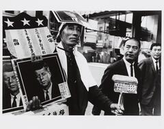 Photograph of a man standing on a sidewalk holding posters of John F. Kennedy.