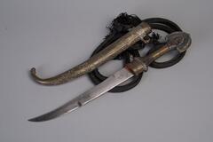 This dagger has a metal blade and an elaborately decorated handle. At the end of the handle is a “peacock tail” pommel. Accompanying the dagger is a silver sheath with a tasseled, black woolen baldric. 