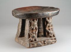 A wooden stool with four posts supporting the seating platform. The posts are decorated with repeating motifs depicting spiders. 