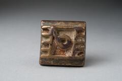 Gold-weight in the shape of a square base with a raised central circle with a short tail surrounded by five raised bars on opposite sides.  