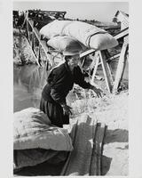 A woman with two long pillows stacked crossed on her head moves mats and bedding materials on the back of a river